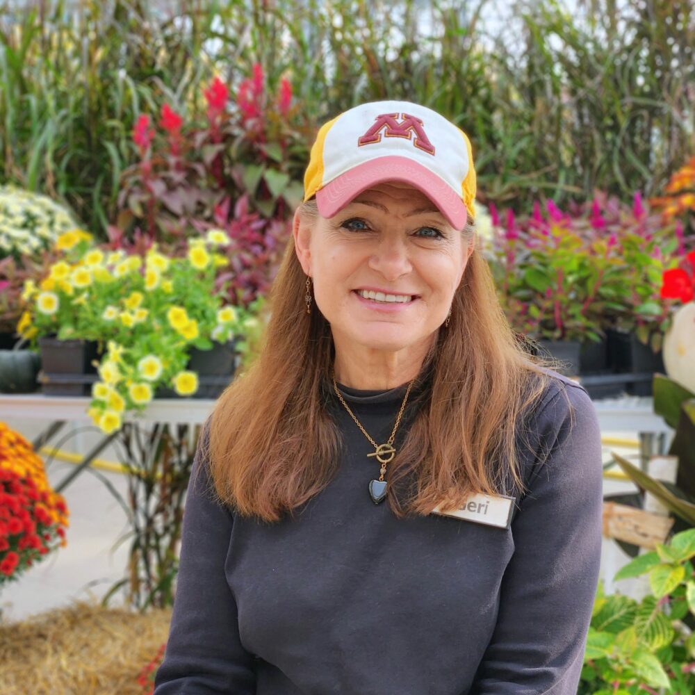 geri vogel sitting in front of fall annuals in the greenhouse wearing a Minnesota hat.